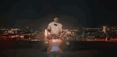 a man sitting on a toilet with a city in the background at night