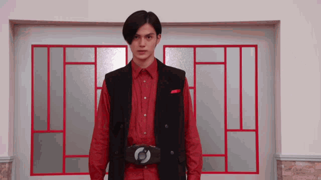 a young man wearing a red shirt and a black vest is standing in front of a window .