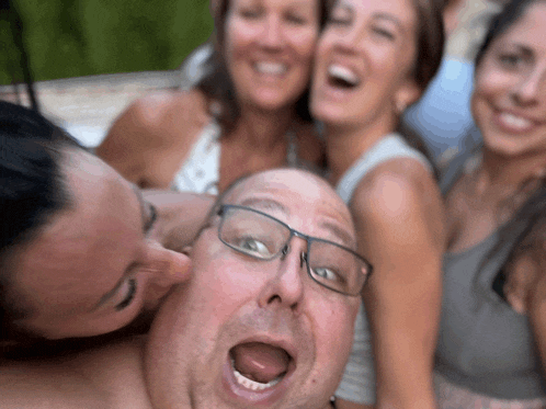 a man with glasses is surrounded by women and one of them is kissing his face