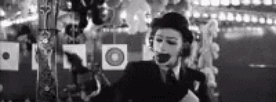 a man in a suit and tie with a mime mask on his face is standing in front of a carousel .