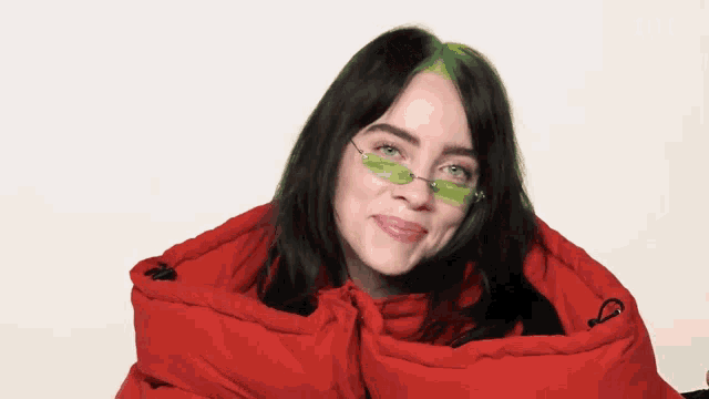 a woman wearing green sunglasses and a red jacket looks at the camera