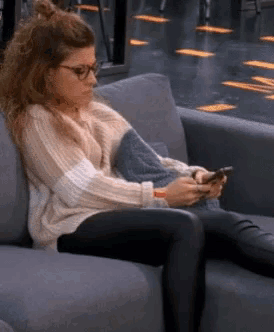 a woman wearing glasses is sitting on a couch looking at her cell phone .