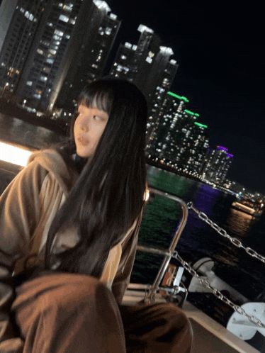 a woman with long black hair is sitting on a boat in front of a city skyline at night