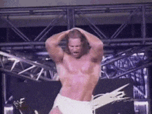a shirtless wrestler in white shorts is standing on a stage with his hands on his head .