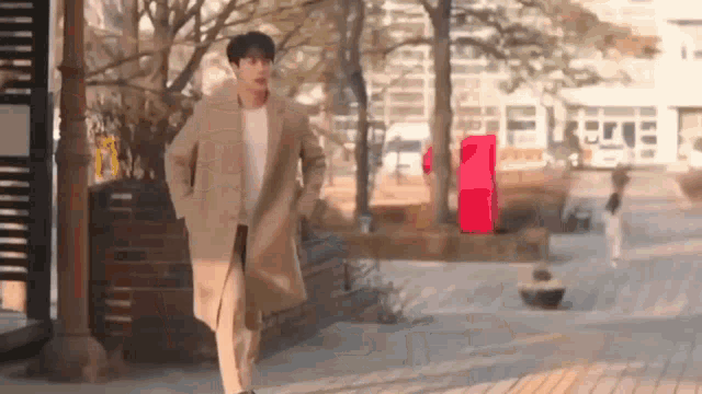 a man in a tan coat is walking down a sidewalk in a park .