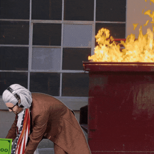 a man wearing headphones is holding a 100 dollar bill in front of a dumpster on fire