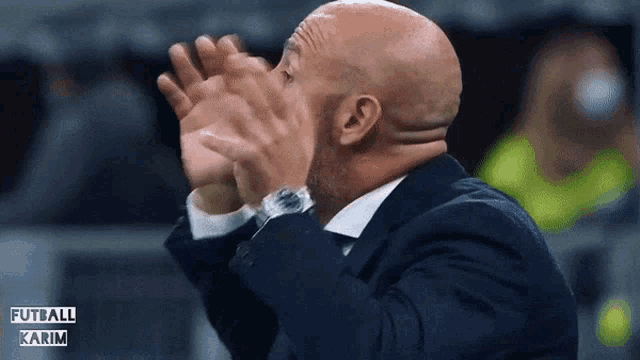a bald man in a suit and tie is clapping his hands in front of a sign that says futball karim