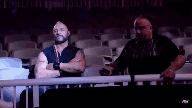 two men sitting in a stadium with aaw on the bottom right