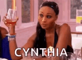 a woman is sitting at a table holding a glass of wine and the name cynthia is on the screen .