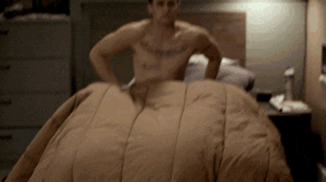 a shirtless man with a tattoo on his chest is standing on a bed with a brown comforter