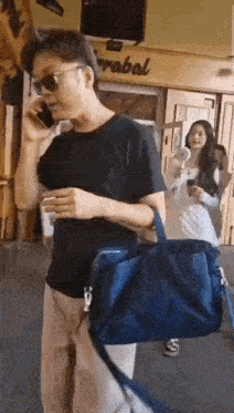 a man is talking on a cell phone while holding a blue bag and a woman is standing behind him .