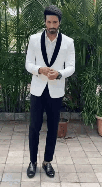 a man in a white tuxedo and black pants is standing on a sidewalk in front of palm trees .