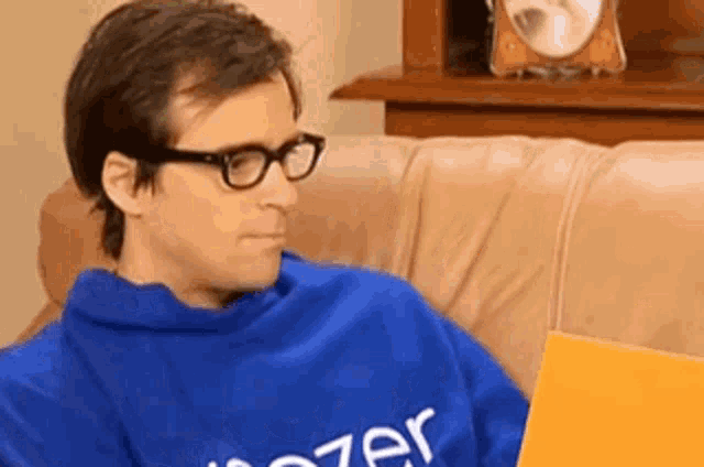 a man wearing glasses and a blue shirt is sitting on a couch holding a book .