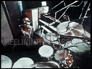 a man singing into a microphone in front of a drum set with the words reelin ' in the background