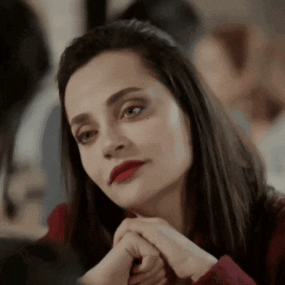 a woman with red lipstick is sitting at a table with her hands folded and looking at the camera .