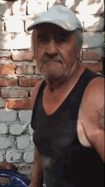 an elderly man wearing a hat and tank top is standing in front of a brick wall .
