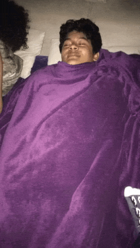 a young man is wrapped in a purple blanket and sleeping on a bed .