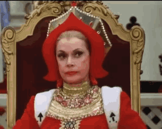 a woman is sitting on a throne wearing a red dress and crown .