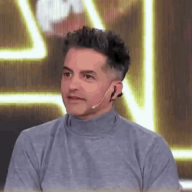 a man wearing a turtleneck sweater and a microphone on his ear is sitting in front of a screen .