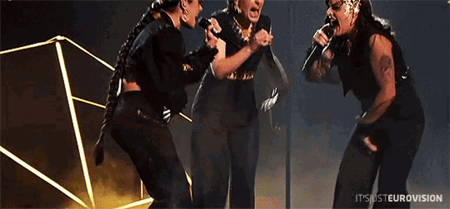 three women singing into microphones on a stage with the words itsjusteurovision below them