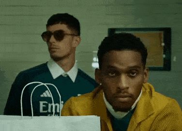 two men are standing next to each other in a room . one of the men is holding a white bag .