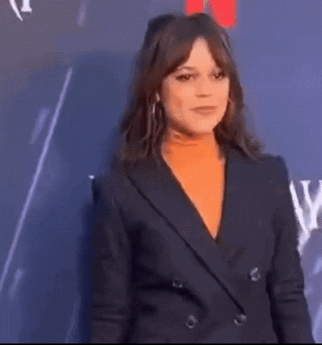 a woman in a black suit and orange turtleneck is standing in front of a blue wall .