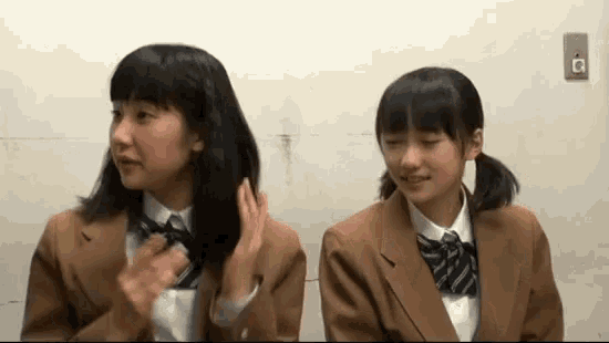 two young girls in school uniforms are sitting next to each other in front of a wall .