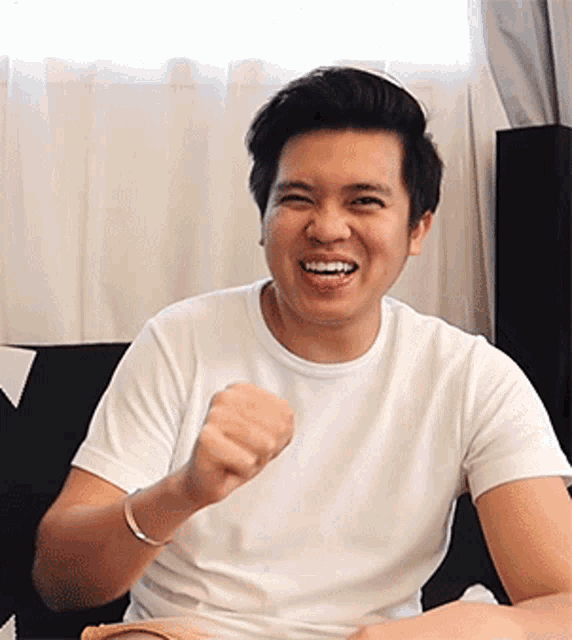 a man in a white t-shirt is sitting on a couch with his fist in the air