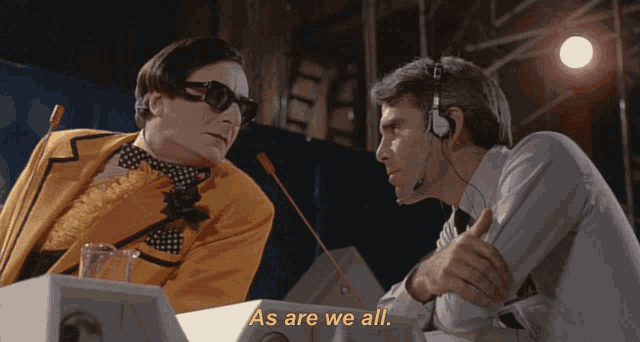 a man wearing headphones talks to another man with the words as are we all behind him