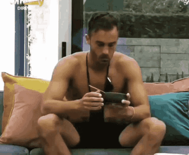 a shirtless man is sitting on a couch looking at a cell phone