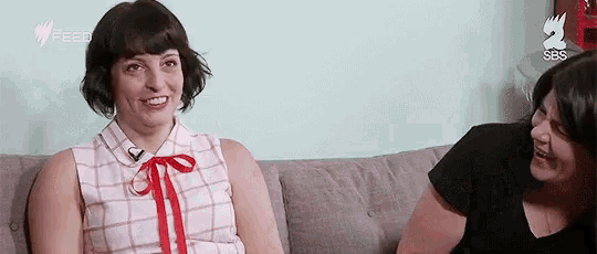 two women are sitting on a couch talking and smiling .