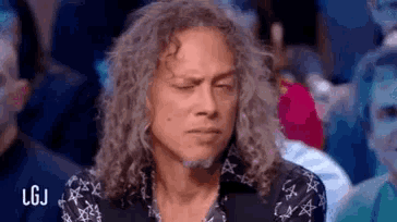 a man with long curly hair and a beard is sitting in front of a crowd with his eyes closed .