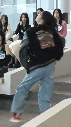 a girl in a black jacket and jeans is dancing in front of a group of girls .