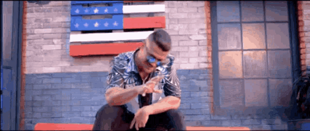 a man wearing sunglasses is sitting on a bench in front of an american flag ..