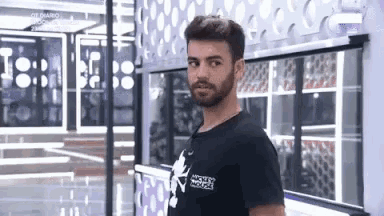 a man with a beard wearing a black t-shirt is standing in front of a window in a room .