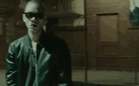 a man wearing sunglasses and a jacket is standing in a dark room .
