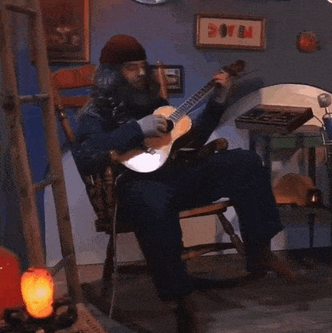 a man sits in a rocking chair playing a guitar with a framed picture on the wall that says ' peace '