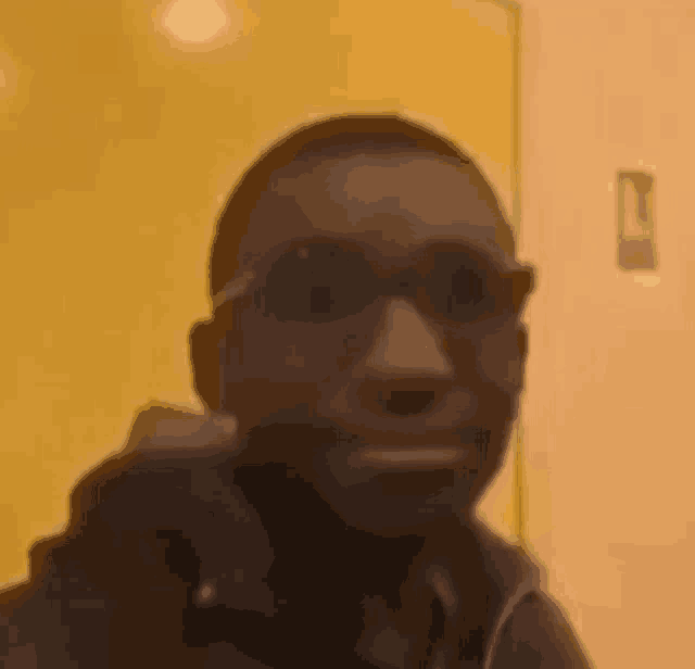 a man wearing sunglasses is making a funny face while sitting in front of a yellow wall .