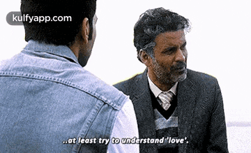 a man in a suit and tie is talking to another man in a denim jacket .