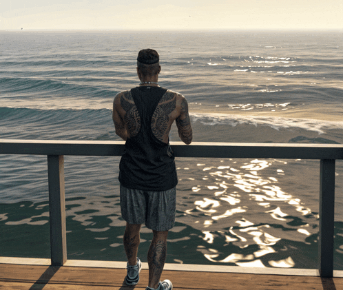 a man with a tattoo on his back stands on a pier looking at the ocean