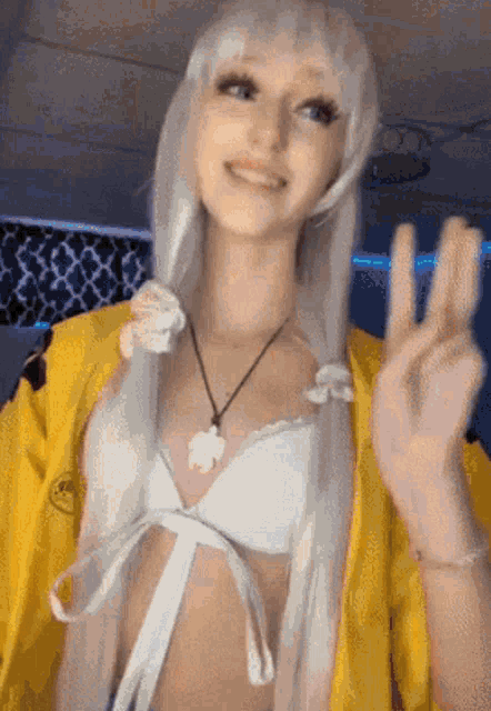 a woman wearing a yellow jacket and a white bikini top is smiling and giving the peace sign .
