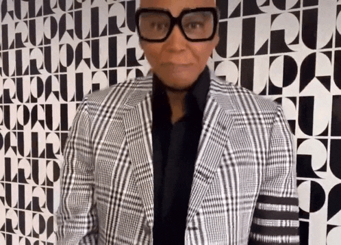 a man wearing glasses and a plaid jacket is standing in front of a wall that has the word vogue on it