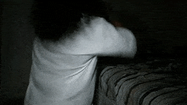 a woman in a white shirt is standing next to a bed in the dark .