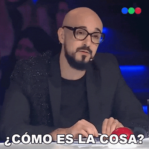 a bald man with glasses and a beard is sitting at a table with the words " como es la cosa " above him