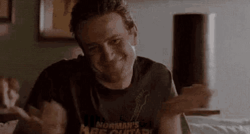 a man in a black t-shirt is sitting on a bed with his hands outstretched and smiling .