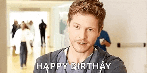a man in a scrub top is standing in a hospital hallway and saying `` happy birthday '' .