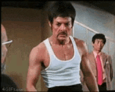 a man in a white tank top with a mustache is standing in a room .
