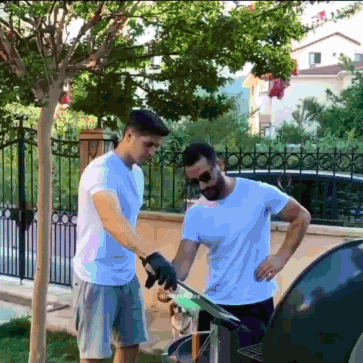two men are cooking on a grill with a fence behind them
