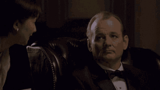 a man in a tuxedo sits in a chair with a woman behind him