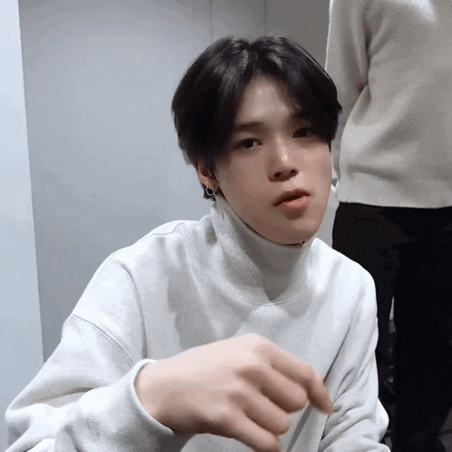 a young man wearing a white sweater with a turtleneck looks at the camera